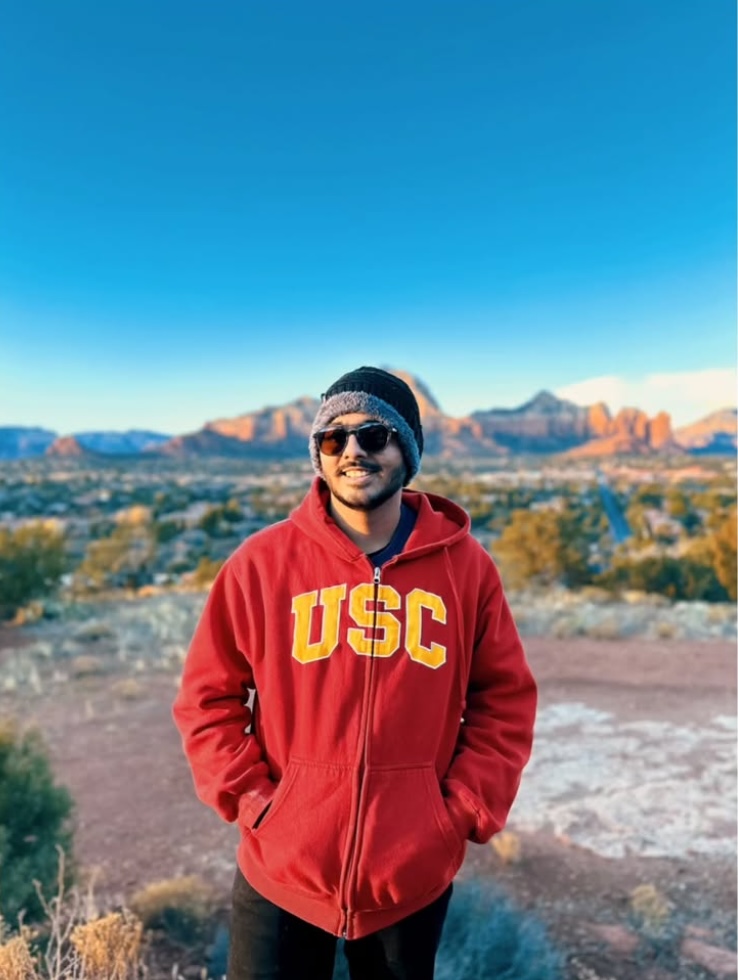 Pathik standing outdoors in a red USC hoodie with a scenic mountain background
